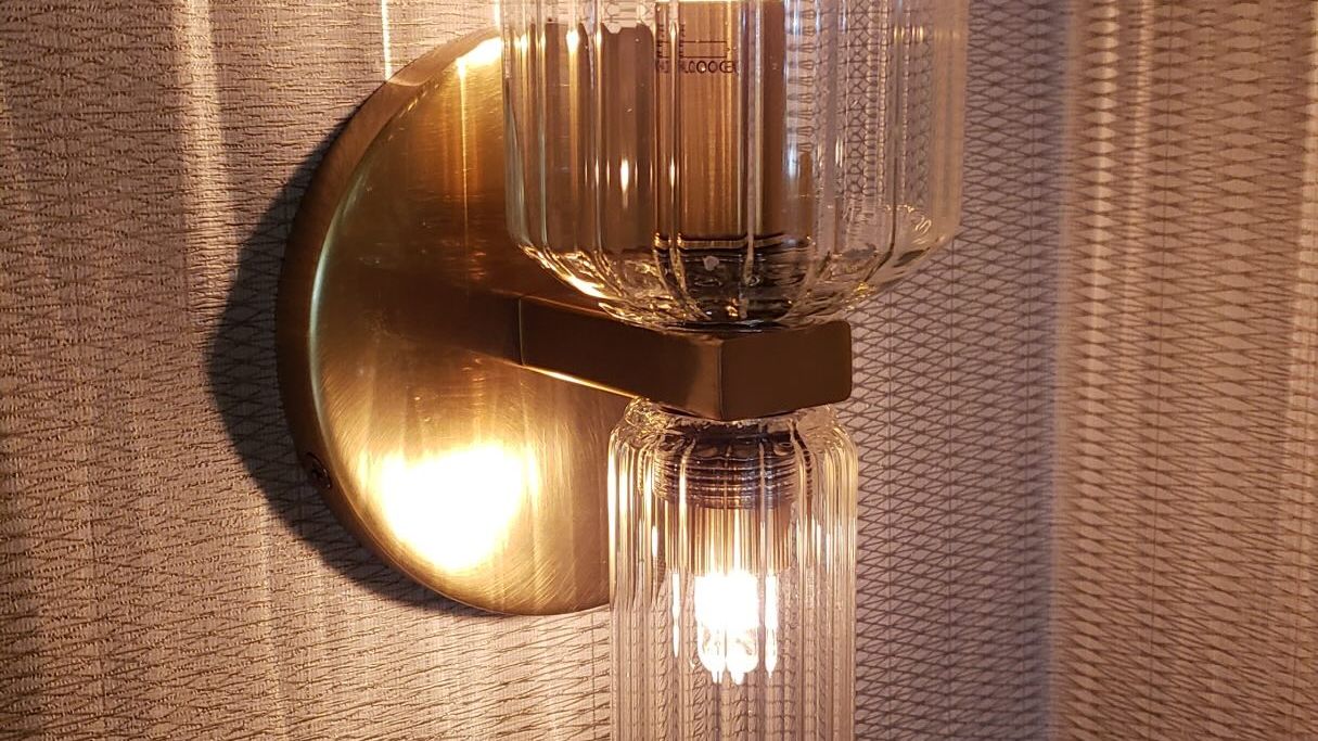 A brass wall sconce with two ribbed glass shades, both bulbs lit, glowing against a pleated wall.