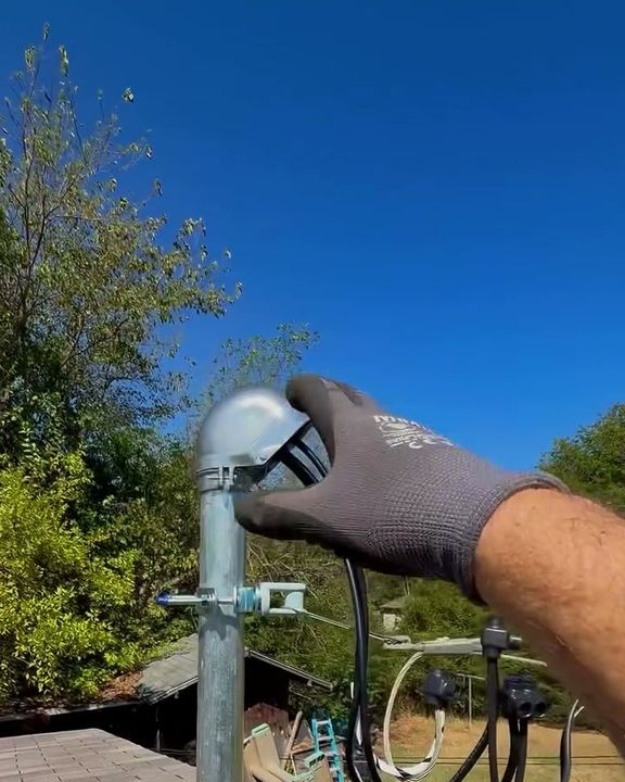 A gloved hand on the weatherhead of a new service riser on a roof, blue sky and trees behind.