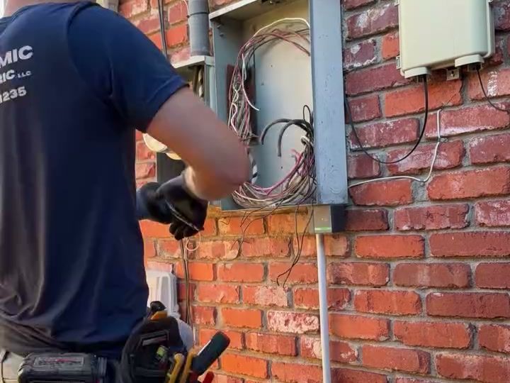 An electrician at the old panel on a red brick wall, its cover off and the old wiring hanging out.