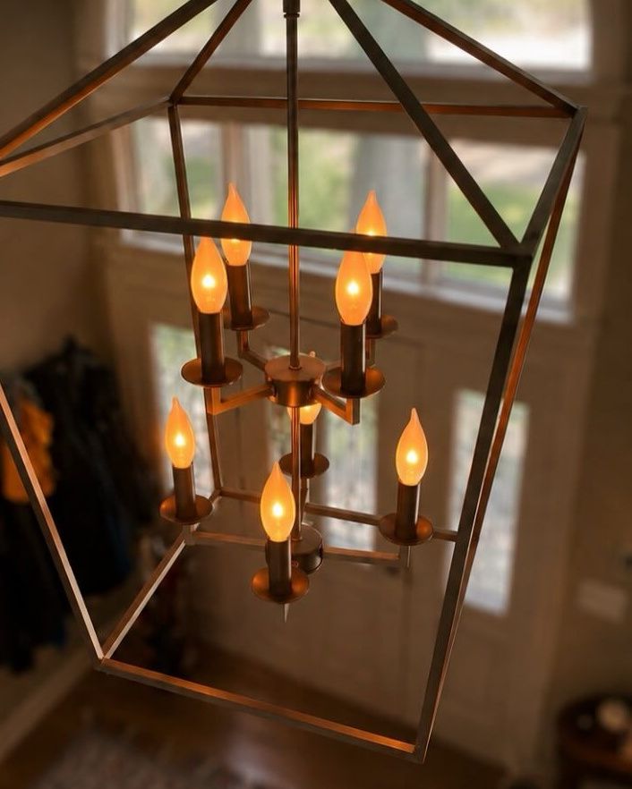 A bronze lantern chandelier with candle bulbs hanging in a bright entryway, an arched window behind it.