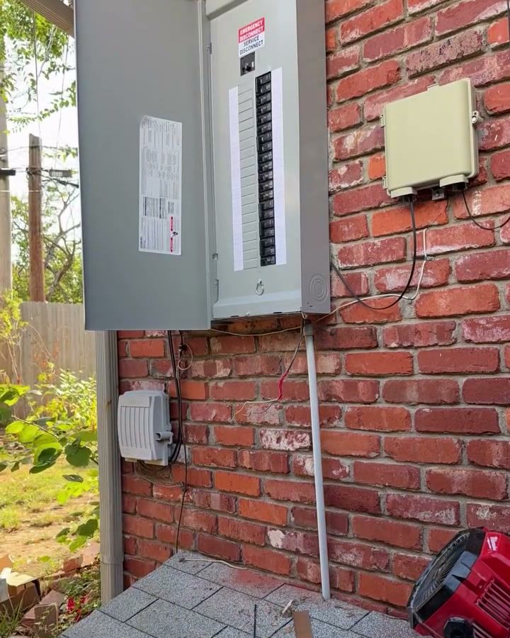 A new grey breaker panel with its door open on a red brick wall, rows of breakers inside, the meter beside it.