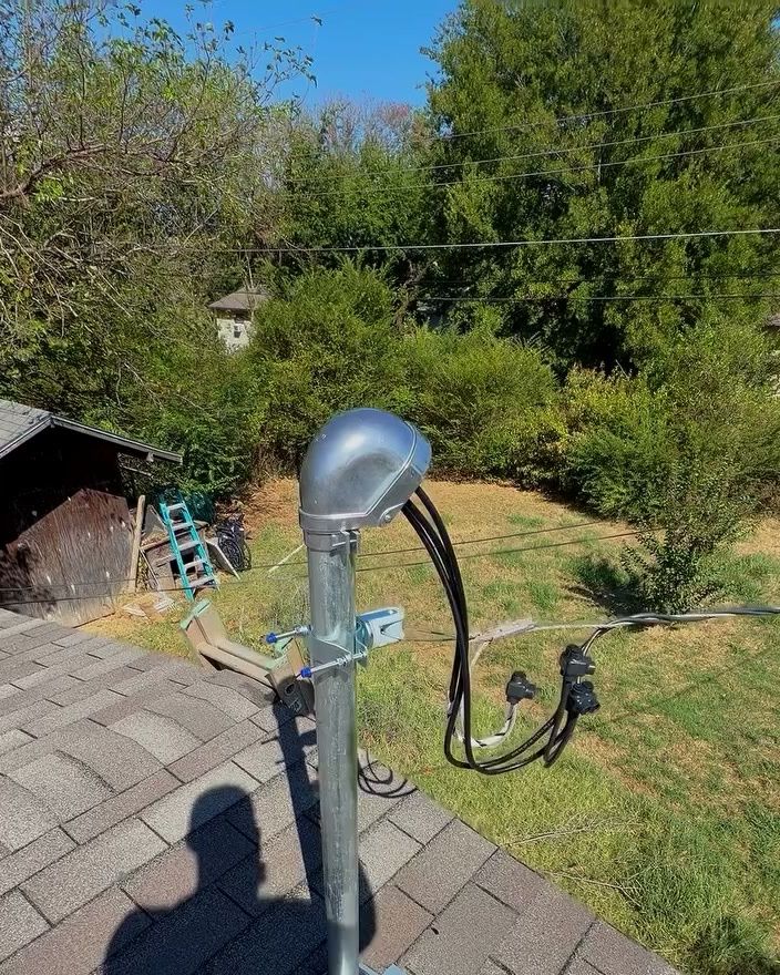 A new galvanized service riser on a shingle roof with its weatherhead and service conductors, a yard and trees beyond.